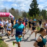 Grupa biegaczy kończy wyścig, przechodząc pod różowym łukiem z napisem META. Tło to zielony las i niebo z chmurami. Uczestnicy noszą kolorowe stroje sportowe.