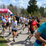 Na zdjęciu grupa biegaczy ubranych w kolorowe koszulki biegnie po asfaltowej drodze. W tle znajduje się las i różowy łuk z napisem META. Jest słoneczny dzień z lekkim zachmurzeniem.