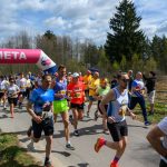 Biegacze przekraczają różowy łuk z napisem META. Mają kolorowe stroje sportowe. Tło to drzewa i niebo z chmurami.