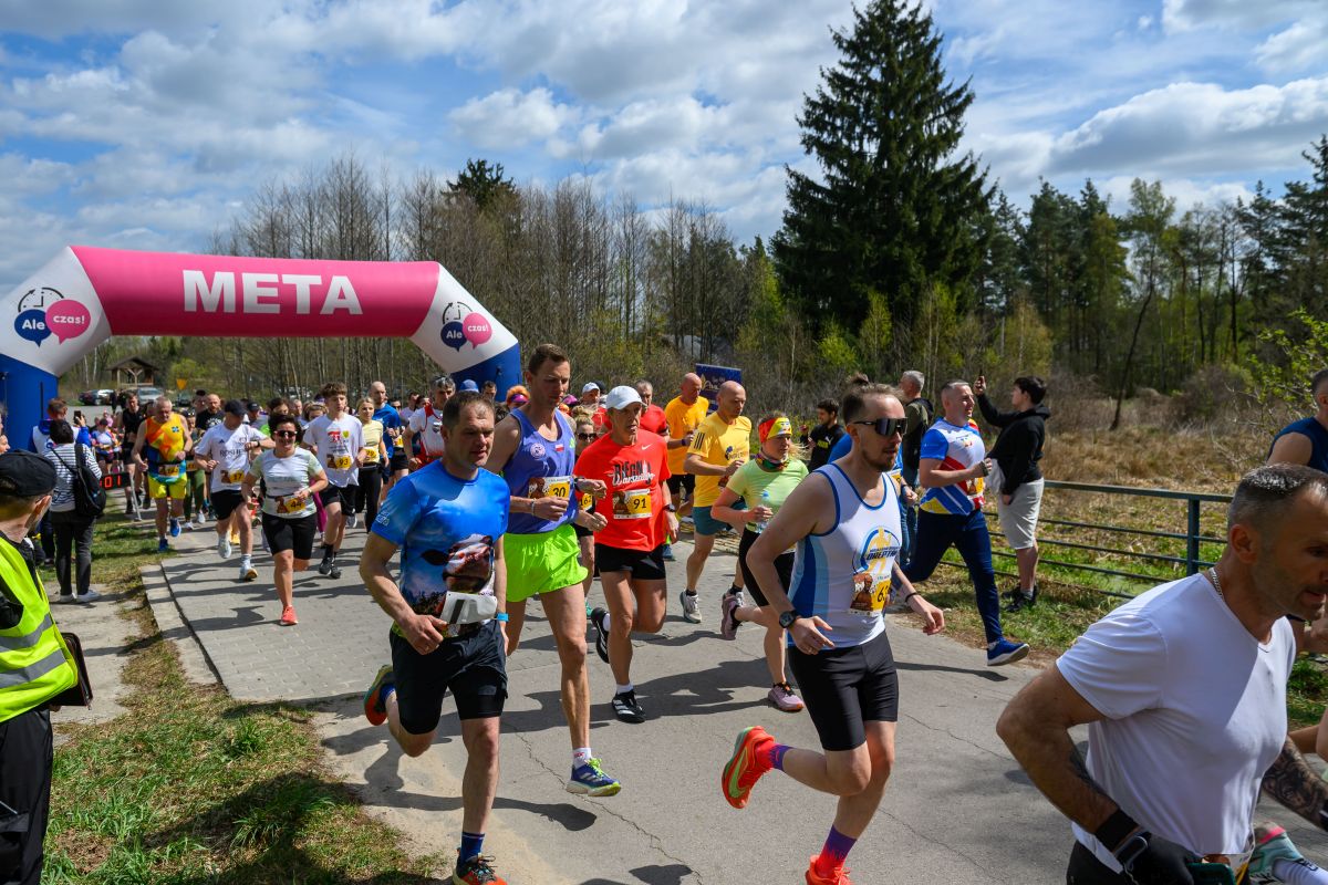 Biegacze przekraczają różowy łuk z napisem META. Mają kolorowe stroje sportowe. Tło to drzewa i niebo z chmurami.