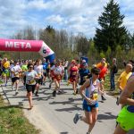 Grupa biegaczy przekracza linię mety na zalesionej drodze. Namiot z różowym napisem META; ludzie w kolorowych strojach. Słoneczny dzień, niebo z chmurami.