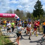 Grupa biegaczy przekracza różowy łuk mety na zewnątrz. W tle drzewa i niebo z chmurami. Atmosfera radości i wysiłku.