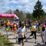 Grupa biegaczy przekracza metę na świeżym powietrzu. Nad nimi różowy łuk z napisem META. Tło to drzewa i pochmurne niebo.