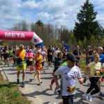 Na zdjęciu widać uczestników biegu na tle lasu, biegających przez linię z napisem META. Ludzie ubrani są w kolorowe stroje sportowe. Pogoda jest słoneczna, a niebo zachmurzone.