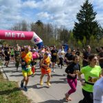 Grupa biegaczy przekracza linię mety pod różowo-niebieskim łukiem z napisem META. W tle widać drzewa i błękitne niebo z chmurami. Uczestnicy noszą kolorowe stroje, tworząc dynamiczną scenę.