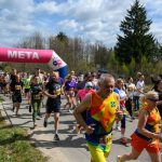 Biegacze w kolorowych strojach przekraczają różowy łuk z napisem META. W tle drzewa i niebo z chmurami. Na poboczu stoi osoba w kamizelce.