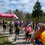 Grupa biegaczy przekracza linię mety oznaczoną różowo-niebieskim łukiem, na tle zieleni i błękitnego nieba.