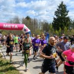 Na zdjęciu grupa biegaczy przekracza linię mety oznaczoną różowym łukiem. Uczestnicy noszą kolorowe stroje. W tle widać drzewa i pochmurne niebo.