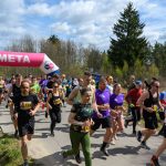 Grupa biegaczy przekracza linię mety oznaczoną różowym banerem. W tle znajdują się drzewa i błękitne niebo. Uczestnicy są ubrani w kolorowe stroje sportowe.