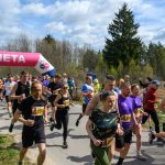 Grupa biegaczy przekracza metę pod różowo-niebieskim balonem. Tło stanowi zielony las. Niebo jest częściowo pochmurne.