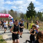 Grupa biegaczy przekracza różowy baner z napisem META na leśnej ścieżce. Słoneczny dzień z niebieskim, chmurzastym niebem. W tle drzewa i kibice.