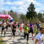 Biegacze przekraczają różowo-białą metę na ścieżce w zielonym parku. Uczestnicy noszą kolorowe stroje sportowe. Słońce świeci, tworząc jasną i pogodną scenerię.