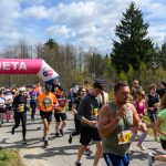 Tłum ludzi biegnie w maratonie, przechodząc przez różowo-białą bramę z napisem META. Uczestnicy noszą kolorowe stroje sportowe. W tle widać drzewa i błękitne niebo z chmurami.