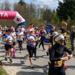 Grupa biegaczy w kolorowych strojach mija różowy balon z napisem Meta. W tle drzewa i niebo. Niektórzy robią zdjęcia.