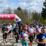 Grupa biegaczy przekracza różowy łuk z napisem META. Ubrani w kolorowe stroje, biegną na tle drzew i nieba. Pogoda jest słoneczna.