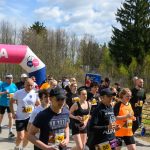 Grupa biegaczy przekracza różowy balon oznaczony META. W tle widać las i niebo z chmurami. Uczestnicy ubrani są w kolorowe stroje sportowe.