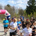 Na zdjęciu widoczna jest grupa biegaczy przy mecie oznaczonej różowym balonem. Tło to zalesiony obszar i niebo z chmurami. Uczestnicy noszą kolorowe stroje sportowe.