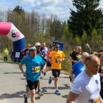 Biegacze przekraczają różowy łuk z napisem META. W tle widać drzewa i niebo z chmurami. Dominują kolory niebieski i zielony.