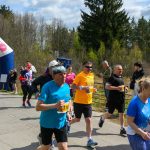 Zdjęcie przedstawia biegaczy na zawodach na tle zielonych drzew. Osoby mają na sobie kolorowe stroje sportowe. W tle widać różowo-białą bramkę startową.