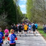 Biegacze na wiejskiej drodze, otoczonej drzewami. Kolorowe koszulki uczestników wyróżniają się na tle zieleni. Słoneczny dzień zachęca do aktywności na świeżym powietrzu.