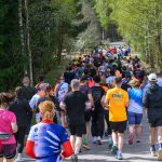 Grupa biegaczy na leśnej ścieżce, otoczonej drzewami. Uczestnicy w kolorowych koszulkach. Słoneczny dzień, zielone tło.