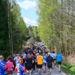 Grupa biegaczy uczestniczy w wyścigu. Są ubrani w kolorowe stroje sportowe. Tło stanowi zalesiona droga i jasno-niebieskie niebo.