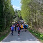 Na zdjęciu grupa biegaczy na wąskiej drodze otoczonej zielonym lasem. Uczestnicy mają kolorowe stroje sportowe. Tło to jasne niebo i drzewa.