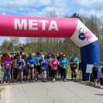 Grupa osób startuje w zawodach za różowo-białym łukiem z napisem META. Tło to drzewa i niebo z chmurami. Uczestnicy są ubrani kolorowo, wokół stoją kibice.