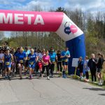 Biegacze przekraczają różowo-niebieski balon z napisem META. Tło to las pod błękitnym niebem. Po bokach stoją kibice.