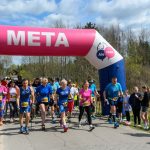 Grupa ludzi startuje w marszu nordic walking pod różowo-białą bramą z napisem META. Uczestnicy noszą kolorowe ubrania sportowe. W tle widać widzów i drzewa, a niebo jest częściowo pochmurne.