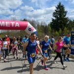 Grupa ludzi z kijkami maszeruje przez różowy łuk z napisem META. W tle drzewa i niebo z chmurami. Widoczny jest baner reklamowy.