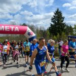 Grupa ludzi kończy marsz nordic walking, przechodząc pod różowo-białym balonem z napisem META. Pogoda jest słoneczna z lekkimi chmurami, wokół widać zielone drzewa. Atmosfera jest żywa i sportowa.