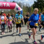 Grupa ludzi w kolorowych strojach kończy wyścig nordic walking pod różowo-białą bramą mety. W tle drzewa i niebo. Z przodu kobieta w niebieskiej kurtce.