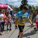 Grupa ludzi maszeruje z kijami do nordic walking pod różowo-niebieskim banerem meta. Noszą kolorowe stroje sportowe. W tle drzewa i niebo z chmurami.