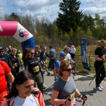 Grupa uczestników nordic walking na zawodach. Tło las i niebo z chmurami. Kolorowe ubrania i różowy łuk mety.