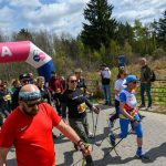 Grupa ludzi z kijkami maszeruje pod różowym banerem oznaczonym META. Mają na sobie kolorowe ubrania sportowe. W tle widać drzewa i niebo z chmurami.