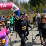 Grupa osób idzie z kijkami do nordic walking pod różowym napisem META. Tło stanowi zielony las i niebo. Uczestnicy noszą kolorowe ubrania.