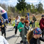 Grupa ludzi uczestniczy w rajdzie nordic walking, mija kolorowy balon startowy. Ciepłe ubrania sugerują chłodny dzień. W tle widać las.