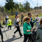 Uczestnicy w sportowych strojach maszerują z kijami nordic walking. Tło stanowi zielone otoczenie z niebieskim niebem. W tle widoczny duży, biało-niebieski balon reklamowy.