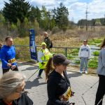 Grupa ludzi uprawia nordic walking na chodniku. Tło to zielony krajobraz z drzewami. Niebo jest jasne i błękitne.