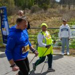 Na zdjęciu widać dwie osoby nordic walking na tle lasu. Ubrani są na sportowo, jedna osoba w niebieskiej kurtce, druga w żółtej kamizelce. W tle są drzewa i młody chłopak w białej bluzie.