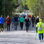 Grupa ludzi, w różnych kolorowych ubraniach, maszeruje po leśnej drodze. Tło stanowią drzewa z zielonymi liśćmi. Część osób trzyma kijki do nordic walking.