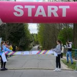 Nad startem, różowy łuk z napisem START. Zawodnicy na drodze w lesie, osoby trzymają taśmę. Tło zielone drzewa.