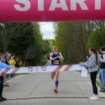 Zawodnik w niebieskiej koszulce przekracza linię mety pod różowym łukiem z napisem START. Wokół są drzewa, a z boku stoi publiczność. Po prawej stronie widoczny jest zegar z czasem.