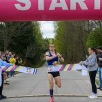 Biegacz w niebieskiej koszulce przecina taśmę na mecie. Nad nim znajduje się różowy nadmuchiwany łuk z napisem START. W tle las i kibice.