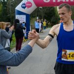 Zawodnik w niebieskiej koszulce kończy bieg i przybija piątkę z kibicem. W tle widać różowy baner START oraz ludzi klaszczących. Scena dzieje się na zalesionej ścieżce.