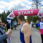 Osoba w niebieskim stroju stoi przed różowym łukiem START, w tle ludzi i drzewa.