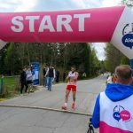 Biegacz w czerwonym stroju przekracza linię startu pod różowym balonowym łukiem z napisem START. Tło stanowi asfaltowa droga otoczona drzewami. W tle widoczni są kibice i uczestnicy.