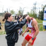 Na zdjęciu kobieta wręcza medal mężczyźnie w stroju sportowym. Tło jest naturalne z drzewami, a wokół kibice. Widzimy również dmuchaną bramę mety.