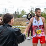 Biegacz w czerwono-białym stroju z medalem, uśmiecha się i podaje rękę kobiecie. W tle osoby obserwujące. Słoneczny dzień w naturze.